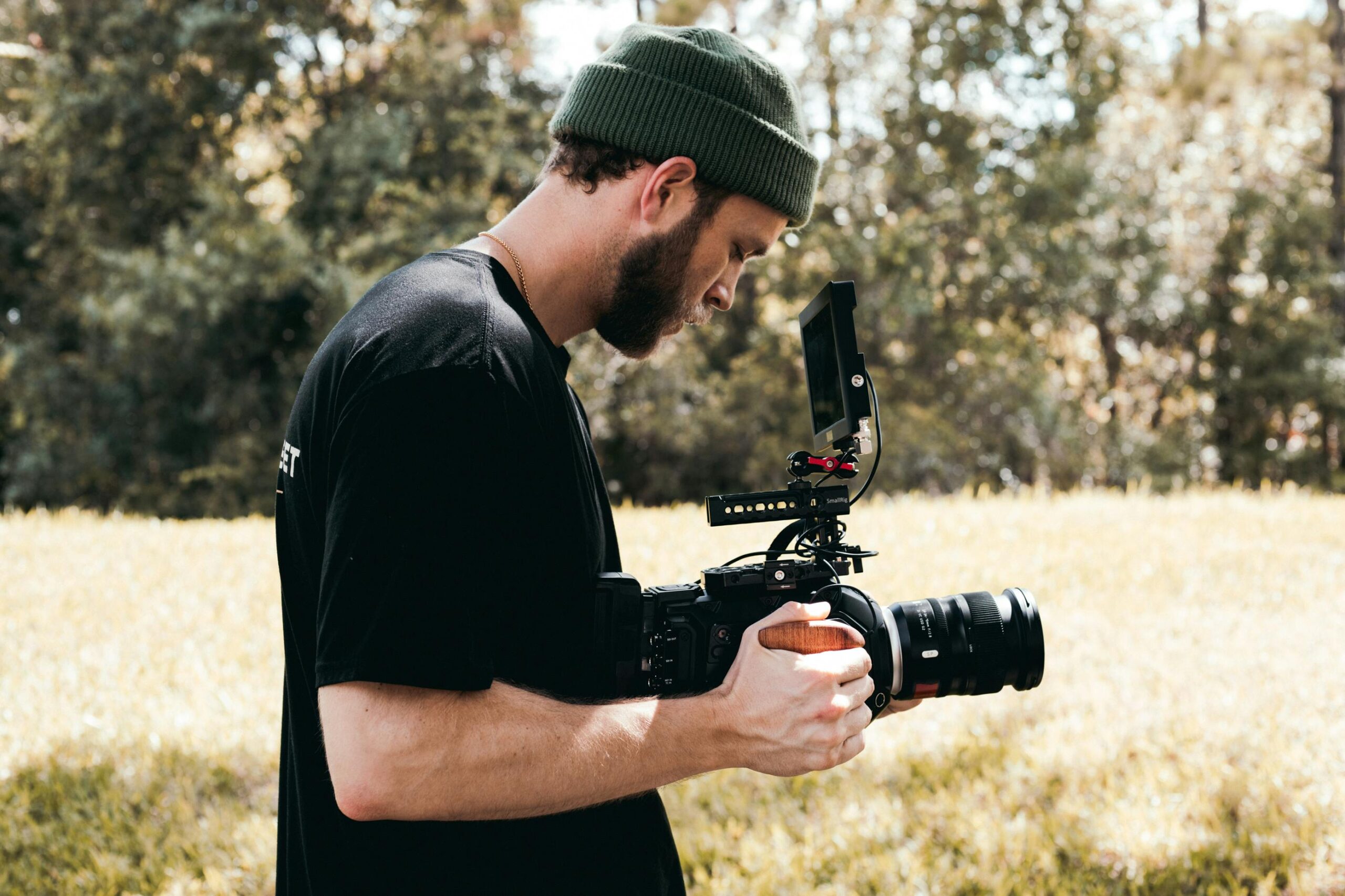 Bearded cameraman wearing beanie filming in grassy field with professional camera gear.